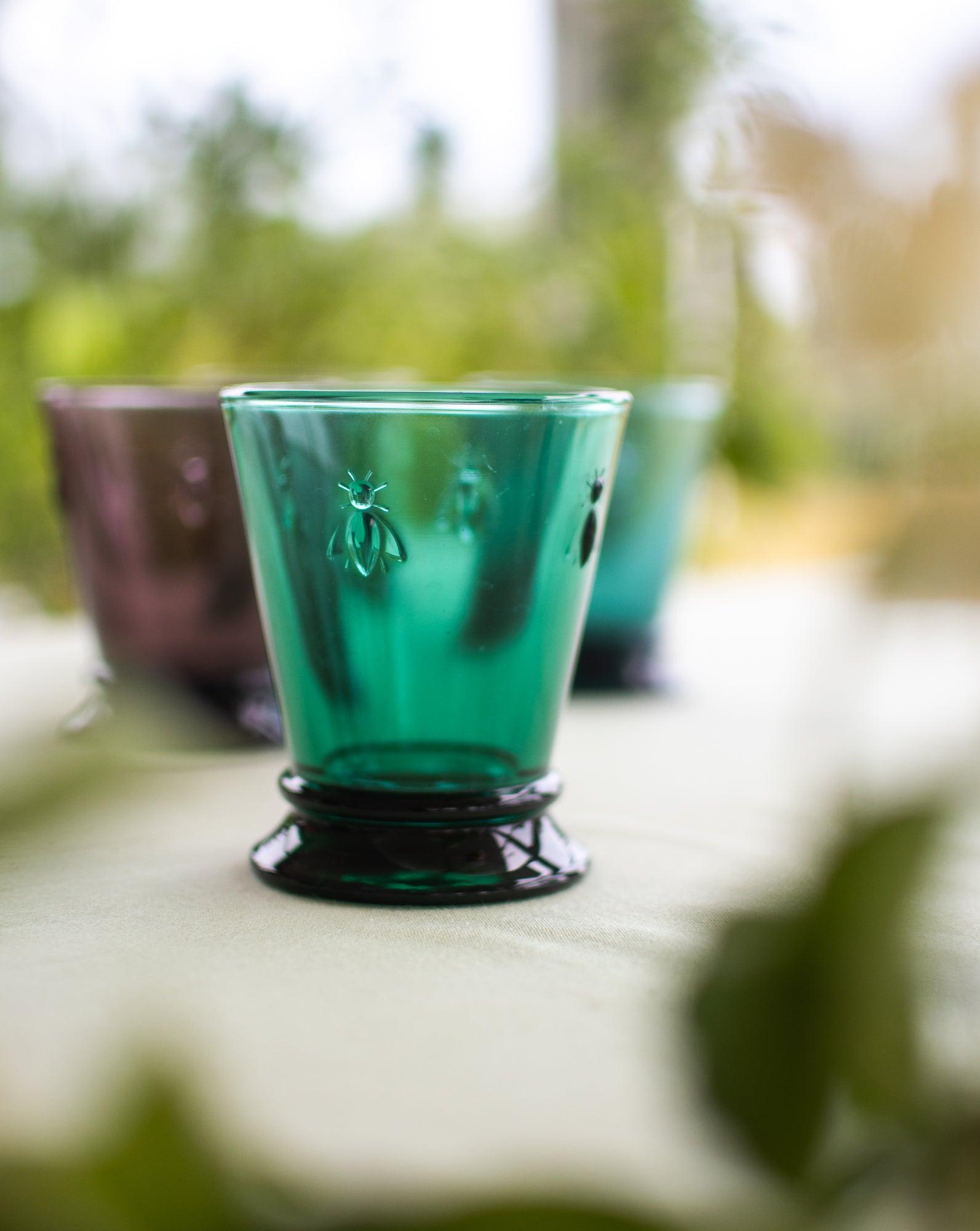 Et vandglas i farven smaragdgrøn stående på et bord med en sløret naturbaggrund og 2 abeille glas i rød og blå