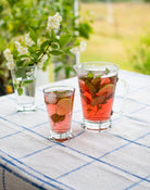 La Rochere glas og kande fyldt med en frisk rød drik med lime og mynte, placeret på et udendørs bord dækket med en blå-hvid dug. En blomsterdekoration er i baggrunden, hvilket skaber en sommerlig og indbydende atmosfære.