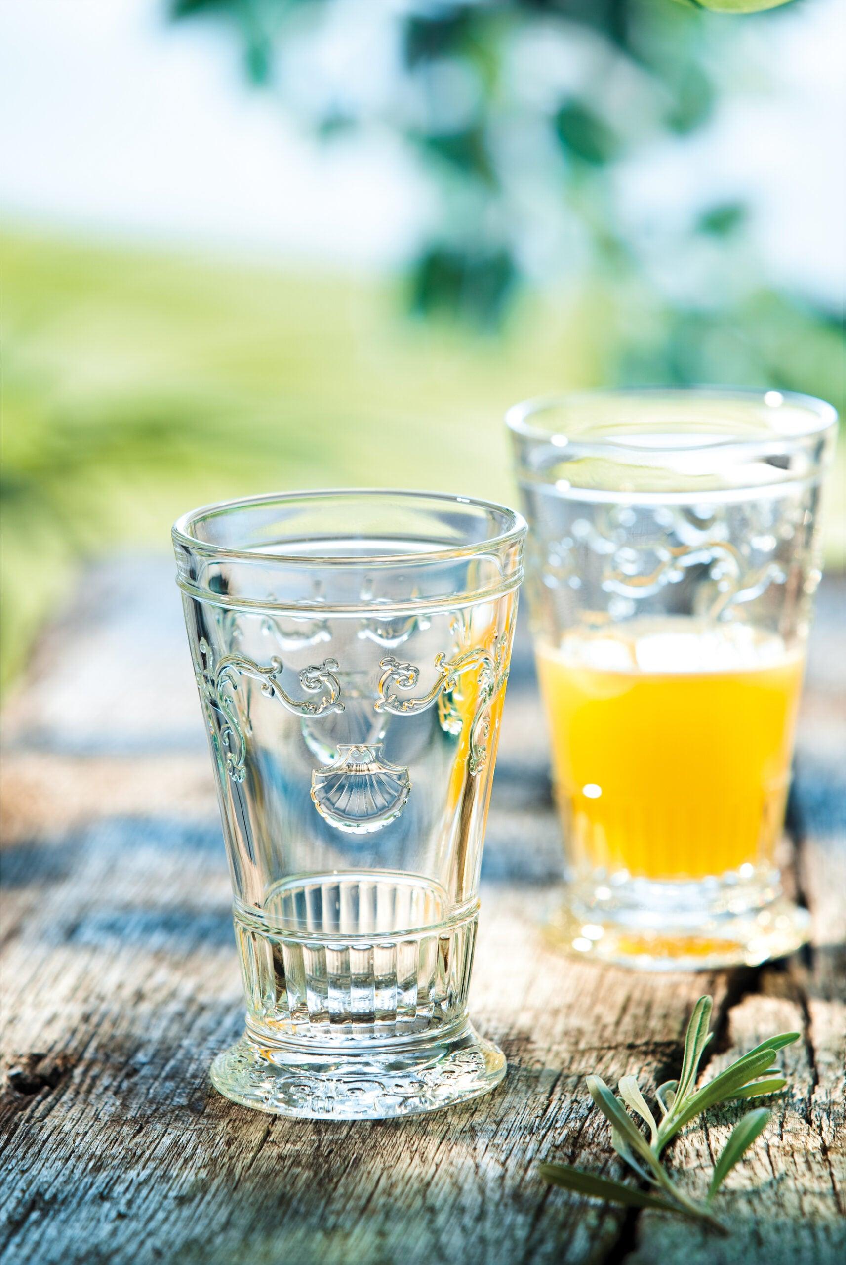 To longdrinkglas stående på en træbjælke med naturen sløret i baggrunden og et fyldt longdrinkglas i serien versailles