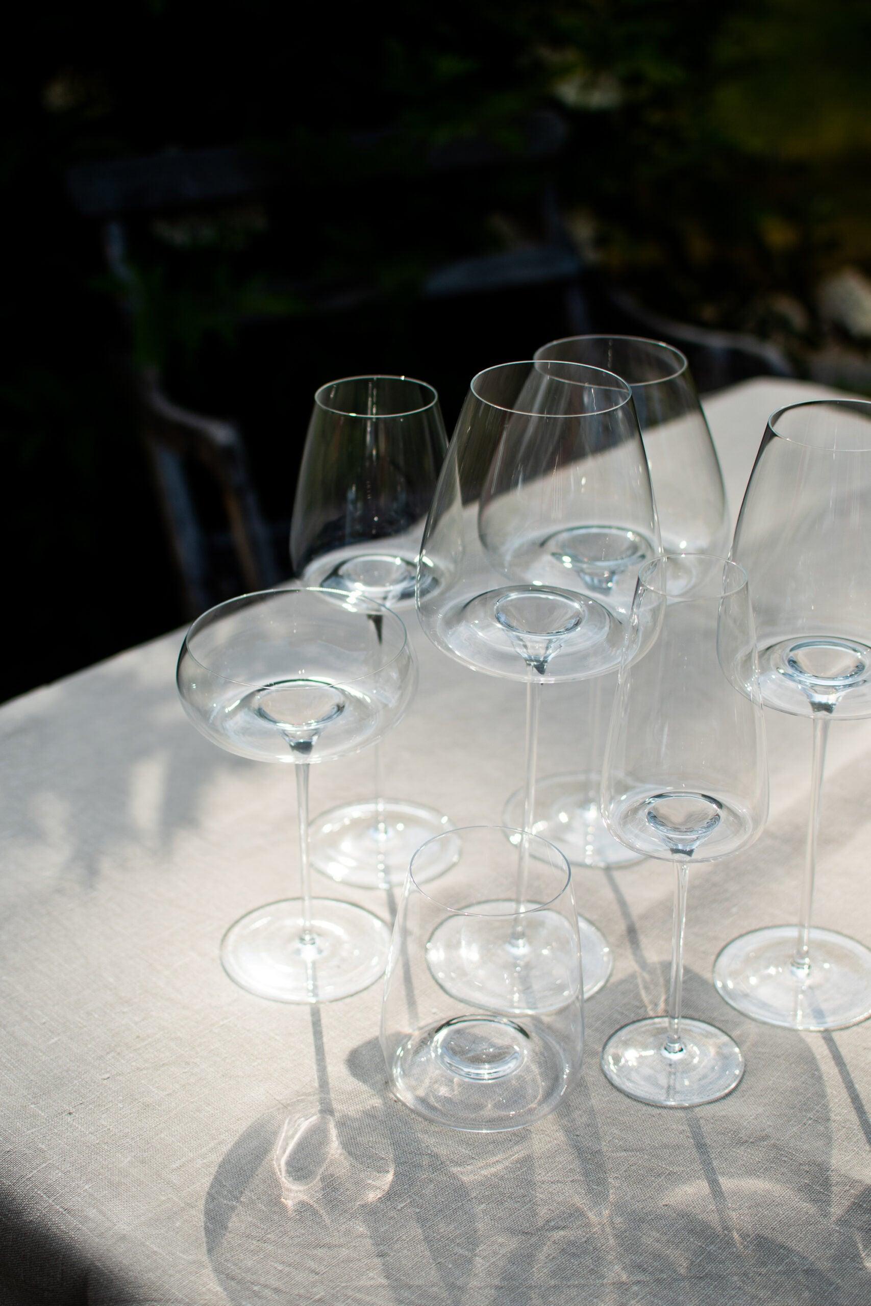 En samling af Zieher glas, inklusive vandglas fra serien Side, står på et bord med en hvid dug i en udendørs setting. Glassene varierer i form og størrelse og er alle fremstillet af mundblæst krystal. Sollyset skaber skygger og fremhæver glassenes klare og elegante design.