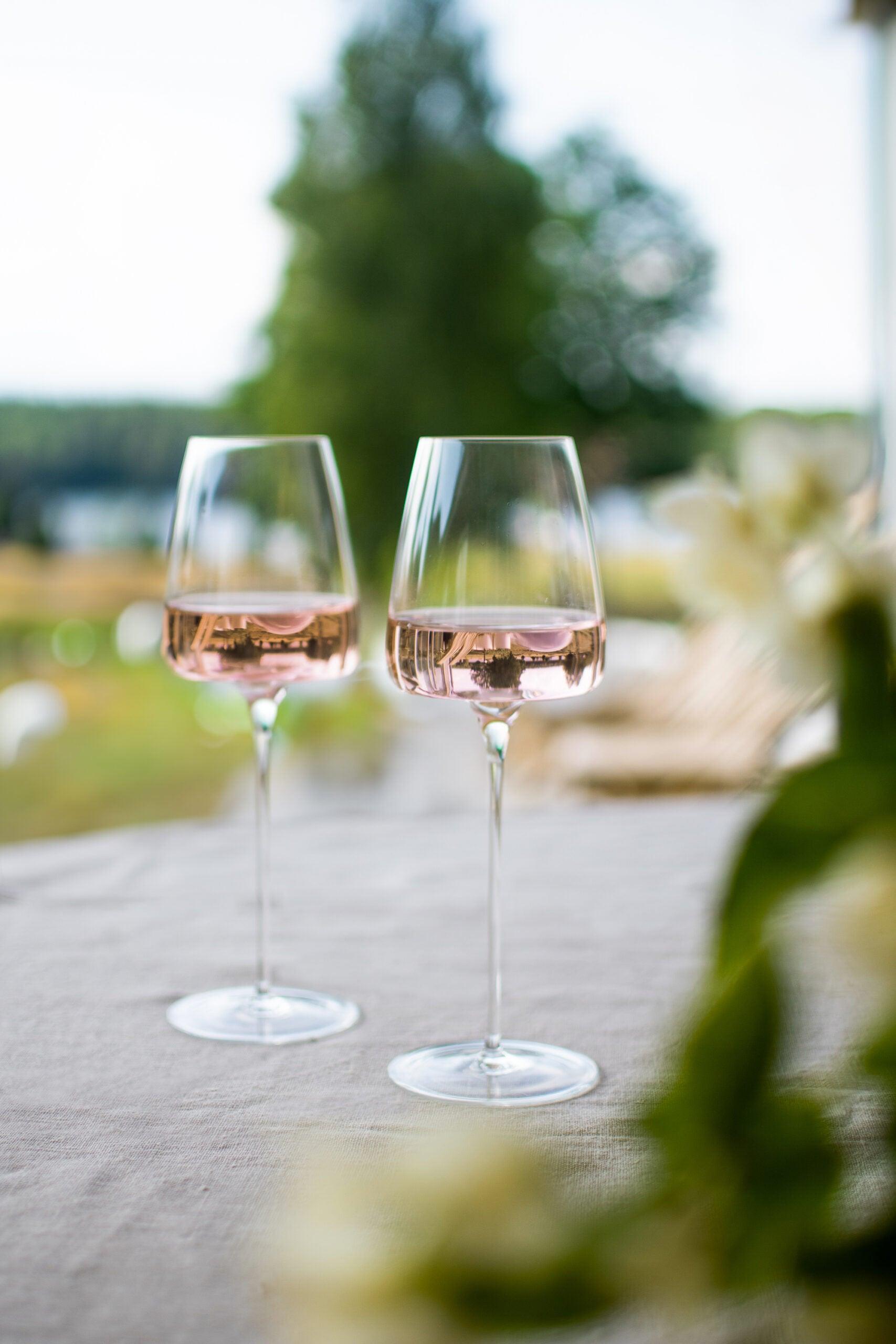 To Zieher Hvidvinsglas fra serien Vision Fresh står på et bord med hvid dug, fyldt med rosévin. I baggrunden ses en sløret udsigt over en frodig have med træer og en sø.