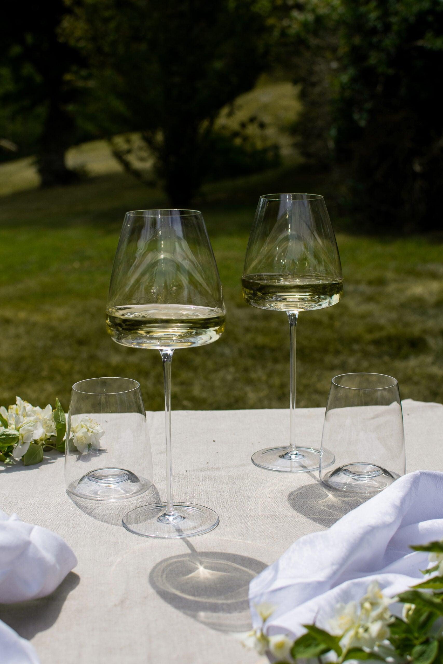 To Zieher Hvidvinsglas fra serien Vision Intense, fyldt med hvidvin, står på et bord med hvid dug i en udendørs setting. Ved siden af glassene ligger hvide blomster og stofservietter. I baggrunden ses en grøn græsplæne med træer.