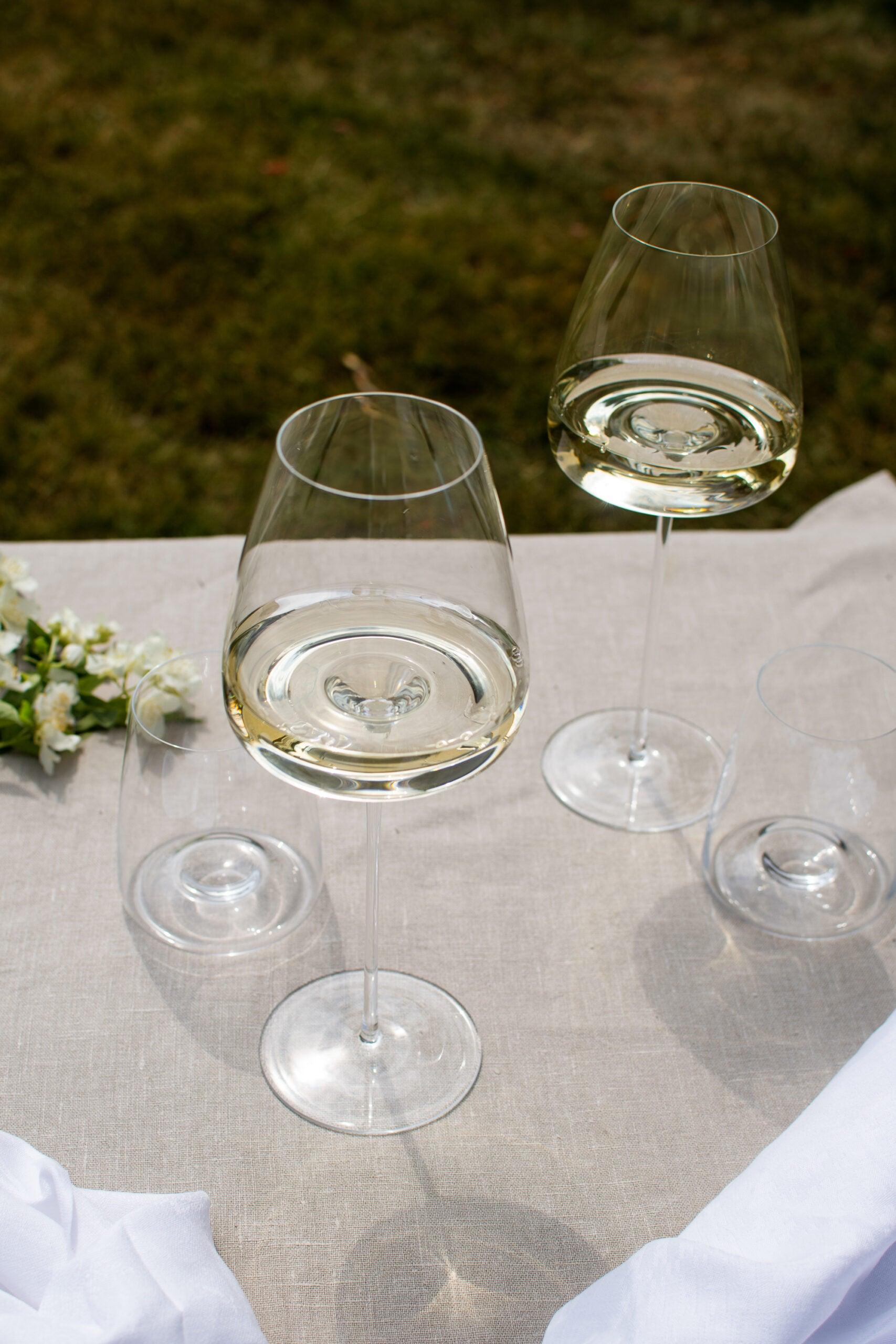 To Zieher Hvidvinsglas fra serien Vision Intense står på et bord med hvid dug, fyldt med hvidvin. Ved siden af glassene ligger hvide blomster. I baggrunden ses en grøn græsplæne.